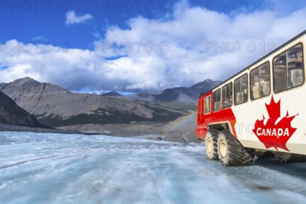Red and white tourist bus driving on the surface of the athabasca glacier in jasper national park, with mountains, cloudy sky and a small rainbow in the background