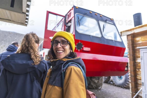 Tourist wearing a yellow beanie and glasses smiles near a large red bus, used to transport visitors onto the athabasca glacier in jasper national park, alberta, canada
