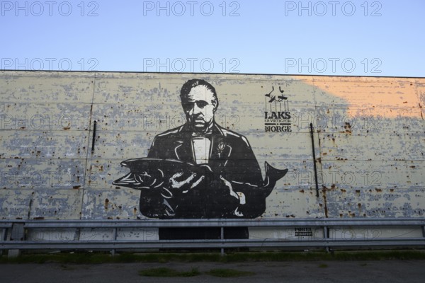 A large mural shows Marlon Brando in the Godfather holding a fish. Mural by street artist Pobel. The inscription means salmon is important for Norway. The art is in black and white and looks rough and urban, Vardø, Finnmark, Norway