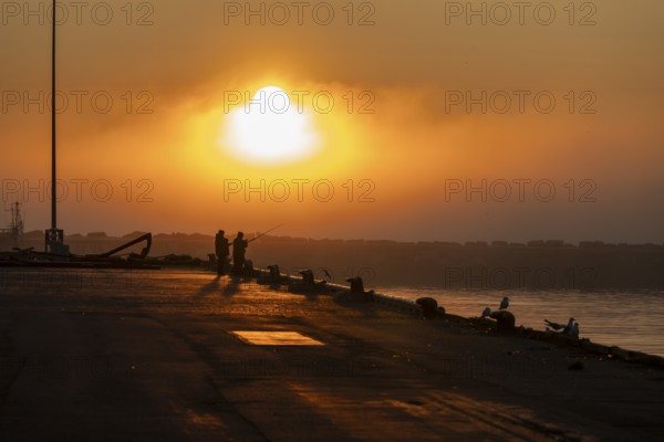 Silhouettes of people fishing at harbour at sunset under a warm orange sky, Vardø, Finnmark, Norway