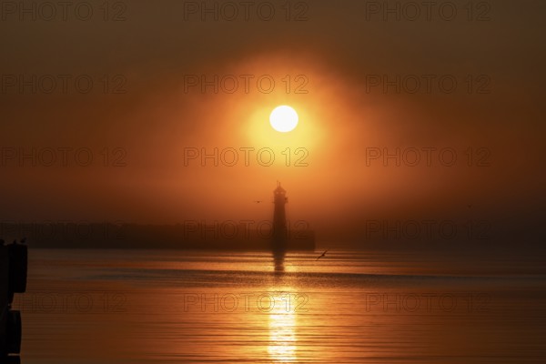 A lighthouse at the Vardö pier stands silhouetted against an orange sunset over the sea reflecting the colors of the sky, Vardø, Finnmark, Norway