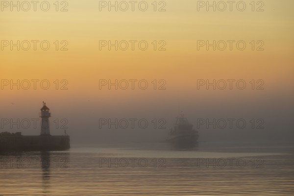 A lighthouse at the entrance to Vardö harbour at sunset with a ship in fog on calm water, Vardø, Finnmark, Norway