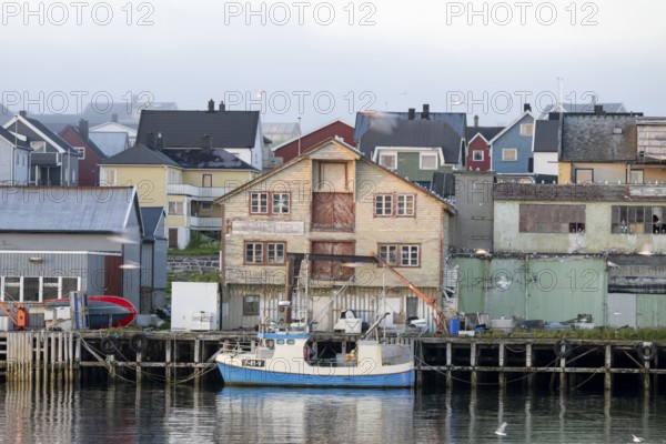 A fishing boat is lying in front of old wooden houses in a quiet harbor, surrounded by morning haze, Vardø, Finnmark, Norway