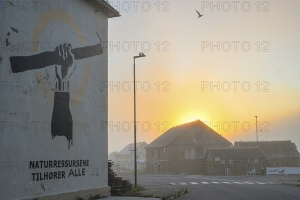Wall painting with stretched arm and gaff. The inscription says Natural resources belong to everyone. Fishing protet on the Barents Sea against fishing quotas. In the background, the Pomor Museum against the midnight sun, Vardø, Finnmark, Norway