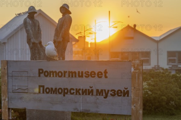 A seagull sits on a museum sign in Norwegian and Russian in front of sculptures and wooden buildings at sunset, Pomor Museum, Vardø, Finnmark, Norway