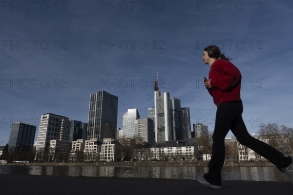 A female jogger walks past the banks of the Main in front of Frankfurt's banking skyline, Mainufer, Frankfurt am Main, Hesse, Germany