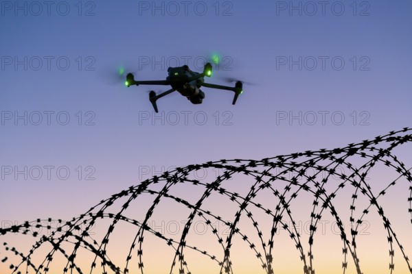 Symbolic picture, illuminated drone flying over barbed wire against the evening sky, airport, threat, espionage