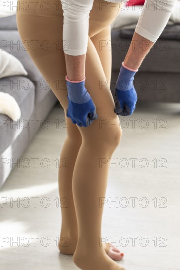 Woman puts on medical compression stockings with special gloves while standing in her home