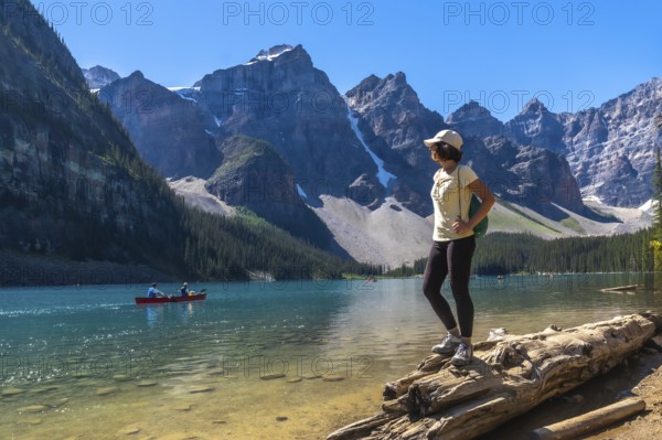 Female tourist standing on a log, admiring turquoise waters of moraine lake with canoers, surrounded by the majestic canadian rockies on a sunny summer day