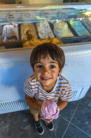 Young boy smiles while holding a scoop of strawberry ice cream in front of a display case filled with various flavors in an ice cream shop in vancouver, british columbia
