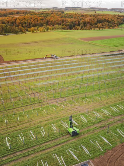 Tractor works in a green field with solar plant construction beginning, surrounded by autumnal forest, energy transition, construction of PV open space, Baden-Württemberg, Germany