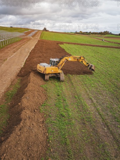 Excavator moves soil over a wide field under cloudy sky, Energiewende, construction of PV open space, Baden-Württemberg, Germany