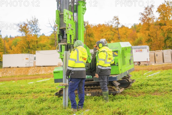 Two workers work with a green machine in front of autumn trees in a meadow, Energiewende, construction of PV open space, Baden-Württemberg, Germany