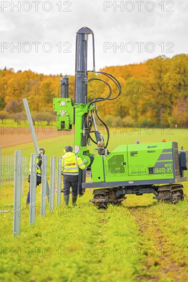 Two workers fix poles with a large, green machine in a field in an autumn landscape, Energiewende, construction of PV open space, Baden-Württemberg, Germany