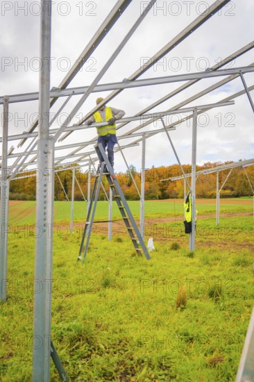 Workers stand on a ladder, assemble metal scaffolding over a green area surrounded by fields, energy transition, construction of PV open space, Baden-Württemberg, Germany