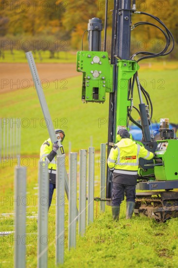 Workers install metal poles in a field with a large green machine in the background, Energiewende, construction of PV open space, Baden-Württemberg, Germany