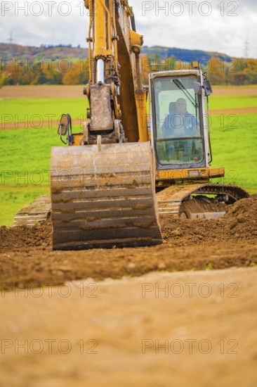 An excavator lifts soil on a field with a green landscape and a hilly background, Energiewende, construction of PV open space, Baden-Württemberg, Germany