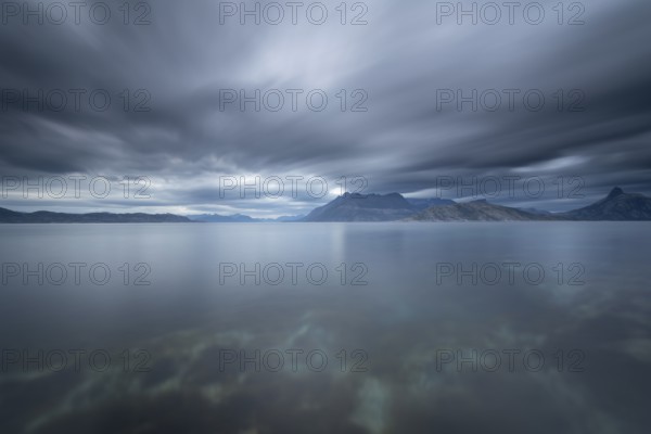 The Vestfjord in Nordland, Norway, near Bodø, impresses with a dramatic atmosphere. Dark clouds sweep across the water and create a mystical atmosphere. The rugged cliffs and majestic mountains are reflected in the tranquil fjord and provide an impressive backdrop