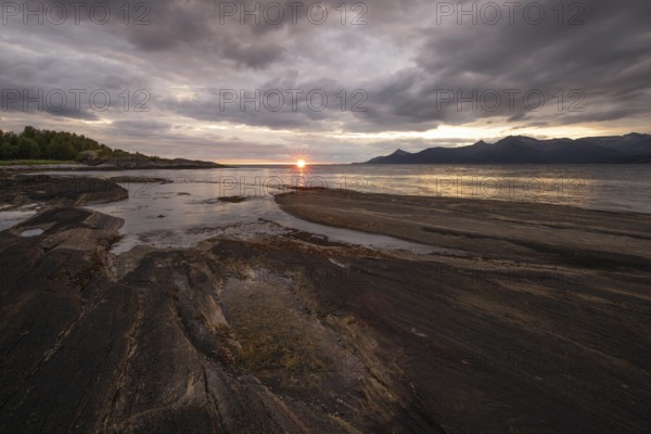 The Vestfjord in Nordland, Norway, offers a dramatic atmosphere near Bodø. Dark clouds are illuminated by the setting sun in shades of red and yellow. The rugged cliffs and mountains are reflected in the quiet fjord, which offers a breathtaking view of Lofoten