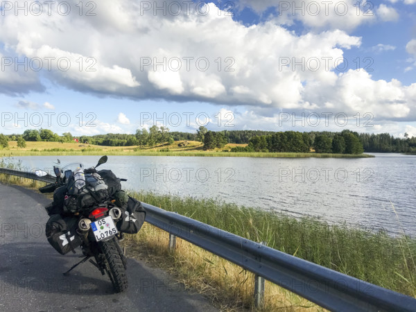 A BMW Sertao Enduro G650GS motorcycle stands on a road next to a lake under a cloudy sky surrounded by green nature, Sparreholm, Södermanslän, Sweden