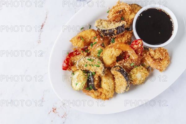 Shrimp and assorted vegetables are deep fried in a light batter and served on a white plate. A small bowl of soy sauce is beside the dish