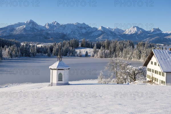 Snowy landscape with chapel, farm and mountains in the background under clear sky, Hegratsrieder See, near Füssen, Ostallgäu, Allgäu, Bavaria, Germany