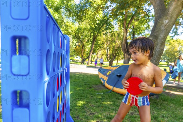 Young boy concentrating on playing a giant connect four game in a park on a summer day, holding a red disc and getting ready to drop it into the grid