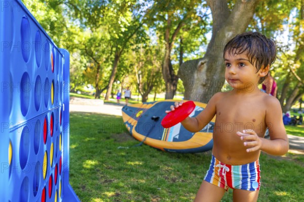 Young child holds a red flying disc, standing near a large connect four game in a park in kamloops, british columbia, enjoying outdoor recreation on a sunny day