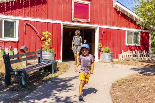 Happy child running from a red barn in the countryside near kamloops, british columbia, canada, with his mother walking behind him on a sunny summer day