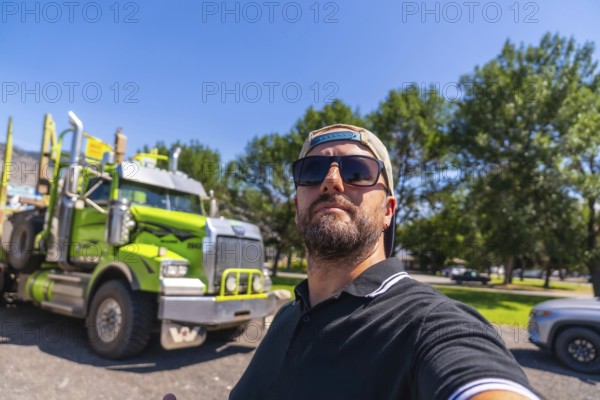 Trucker wearing sunglasses and baseball cap taking selfie with his logging truck in a parking lot in kamloops, british columbia, canada, during a sunny summer day