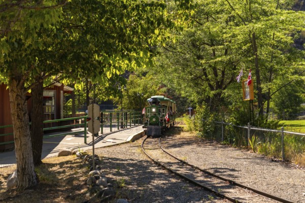 Green and yellow miniature train approaching the station in a sunny summer day in kamloops, british columbia, with canadian flags waving in the background
