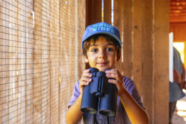 Curious child in a blue cap with good vibes holding binoculars, exploring nature and observing wildlife from a wooden bird hide in kamloops, british columbia