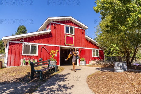 Mother carrying her young daughter as they are leaving a vibrant red barn at the british columbia wildlife park in kamloops, under a clear blue sky