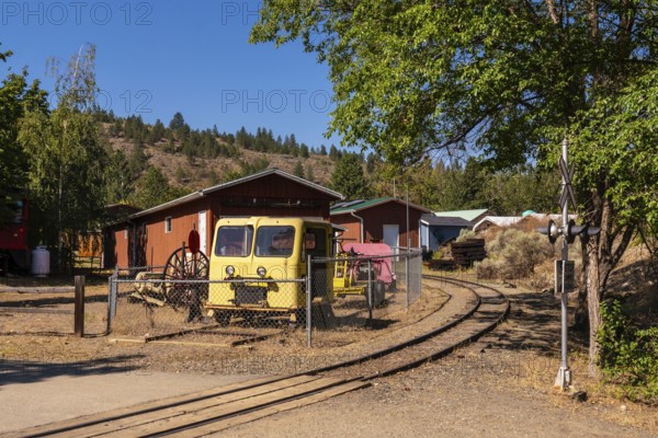 Old yellow locomotive and railway maintenance vehicles resting on a curved rail track in a kamloops yard, surrounded by wooden sheds and forested hills on a sunny day