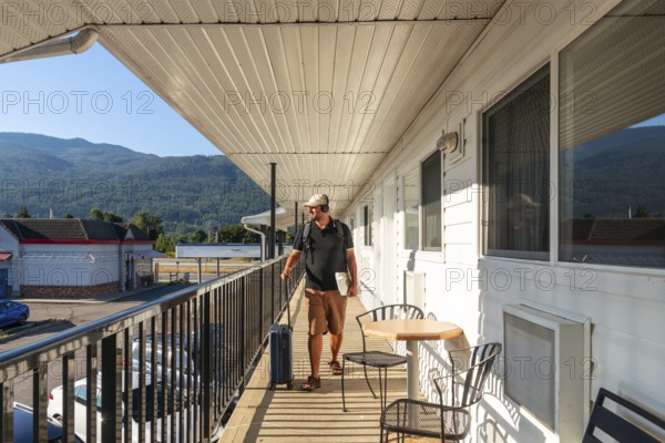 Male tourist wearing headphones and carrying a laptop strolls along a motel balcony, pulling luggage while soaking in the breathtaking mountain view during a peaceful early morning