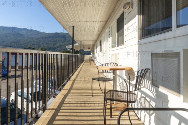 Metal chairs and a small table sit on a motel balcony overlooking a parking lot and a mountain range, providing a relaxing spot to enjoy the view