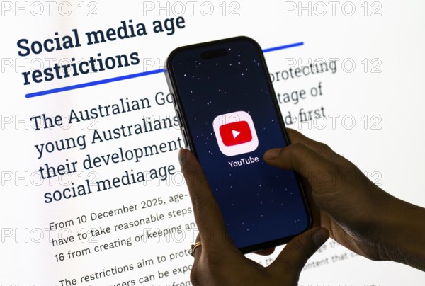 In this photo illustration, a person holds a phone displaying the YouTube app in front of a screen showing information about Australia's new social media age restrictions, in New Delhi, India on 12 December 2025