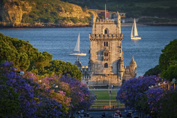 Scenic view of Belem Tower in Lisbon, Portugal, seen over a street with blooming purple jacaranda flower trees street with tourist sailboats on the Tagus River on sunset. Portugal
