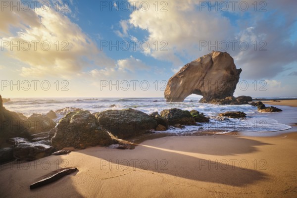 Penedo do Guincho, a large boulder rock arch at Praia da Santa Cruz, Portugal, with ocean waves and sandy beach on sunset