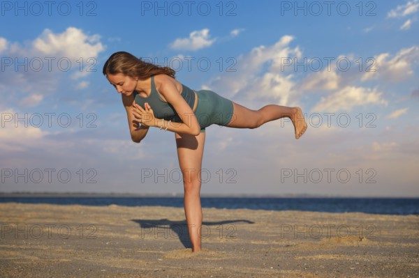 A woman performs Virabhadrasana Warrior 3 pose on the beach during sunset. Her strong stance and focus highlight the beauty of yoga against a tranquil seaside backdrop