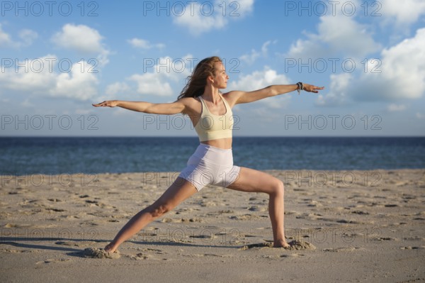 A woman performs the Virabhadrasana 2 Warrior 2 pose on a sandy beach, backlit by a beautiful sunset, enhancing her peaceful yoga practice