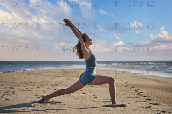 A woman performs Virabhadrasana 1, or Warrior 1 pose, on the beach during sunset. She stands strong against the backdrop of the ocean and colorful sky, embracing tranquility and strength