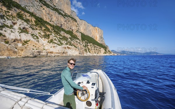 Young man rides a motorboat along the picturesque rocky coast, cliffs and blue sea, Golfo di Orosei, Baunei, Sardinia, Italy