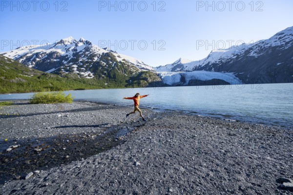 Young man jumping on a pebble beach on a glacial lake, Snowy Mountains and Portage Glacier on Portage Lake glacial lake, Chugach National Forest, Alaska, USA