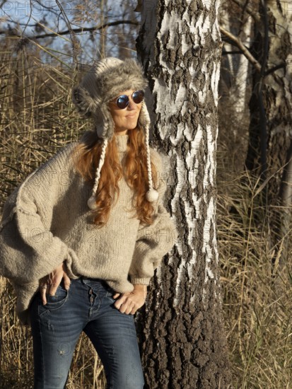 Slender pretty woman with fur hat and pompoms, red hair and warm winter colors between birch forest and reeds