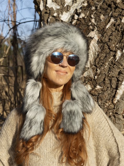 Slender pretty woman with Mongol hat, red hair and warm winter colors between birch forest and reeds