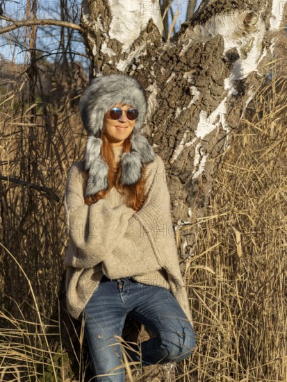 Slim woman with fur hat and sunglasses, red hair and warm winter colors between birch forest and reeds