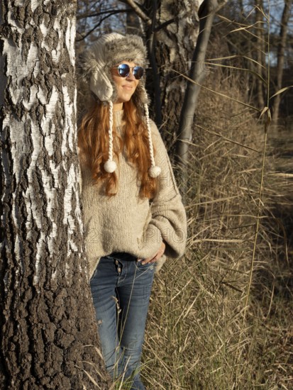 Slender pretty woman with fur hat and sweater, red hair and warm winter colors between birch forest and reeds