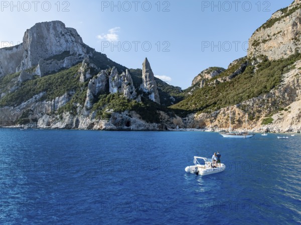 Couple on motorboat off picturesque rocky coast, cliffs with L'Aguglia pinnacle, blue sea and Cala Goloritzé beach, aerial view, Golfo di Orosei, Baunei, Sardinia, Italy