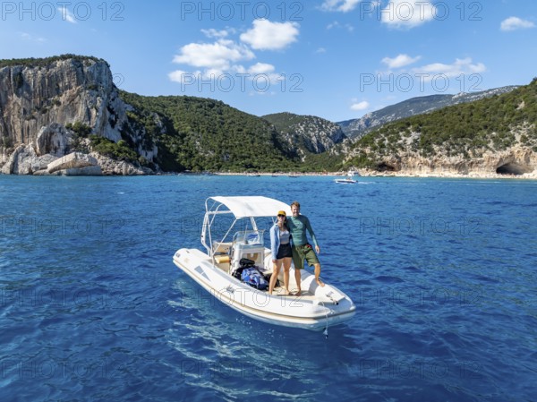 Couple, young woman and young man on motor boat off picturesque rocky coast, cliffs and Cala Luna beach, aerial view, Golfo di Orosei, Baunei, Sardinia, Italy